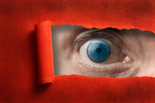 istock Surprised blue eyes looking through from a ripped hole in red paper 985442458