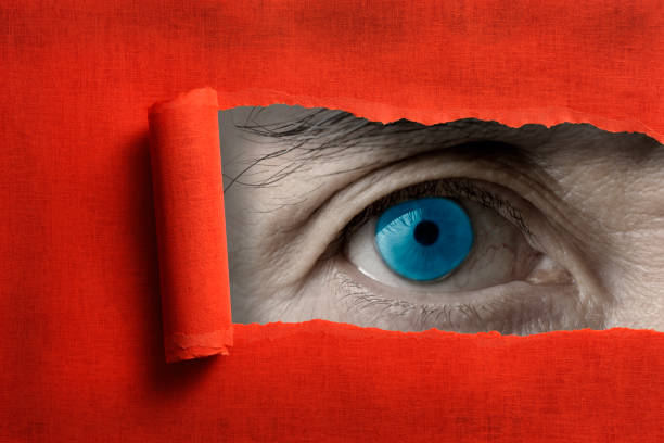Extreme close-up of blue eyes looking through from a ripped hole in red paper.