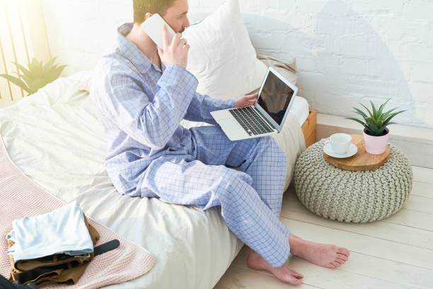 istock Serious confident handsome young entrepreneur in blue pajamas sitting on bed and using laptop while talking on mobile phone in bedroom 985469254