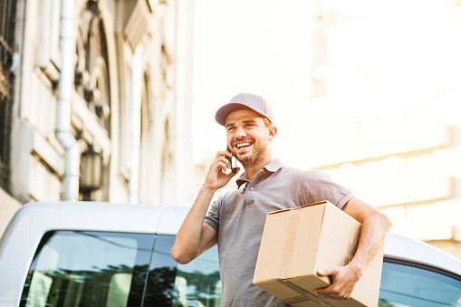 your shipping is here. delivery man in grey shirt with cap standing with his cardboard box on the street calling the client to deliver the package and looking to side