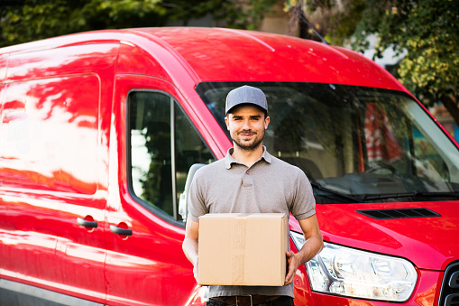 your shipping is here. happy delivery man in grey shirt with cap standing with his cardboard box on the street looking to camera with smile, in front of his delivery car