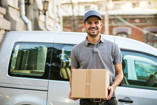 your shipping is here. happy delivery man in grey shirt with cap standing with his cardboard box on the street looking to camera with smile, in front of his delivery car