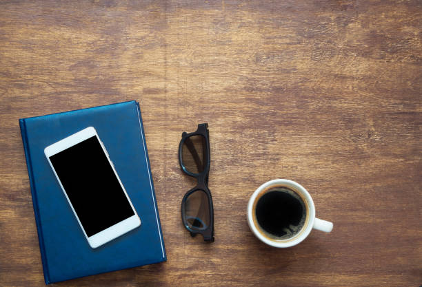 istock Smartphone with black blank space on the screen, blue notebook, black eyeglasses and coffee on wooden background. View from above 985592702