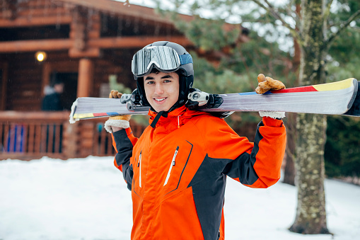 A mixed race teenage boy poses for a portrait in ski wear. He is standing in front of a log cabin.