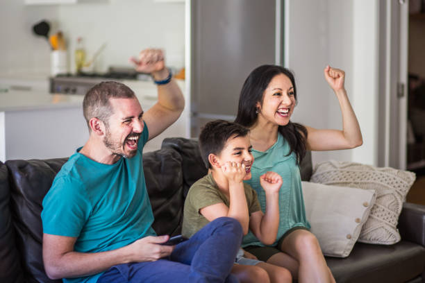 Family cheering while watching TV on sofa Multi-ethnic family cheering while watching TV at home. Enthusiastic fans are sitting on sofa. They are in living room. asian kids watching tv stock pictures, royalty-free photos & images
