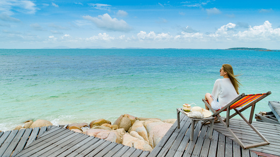 Women wear a sea hat she is happy and sitting on wood comfortable chair coconut drink on the wood bridge and look to beach seaside, cloud and blue sky is endless background, copy space.