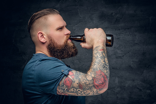 Brutal bearded male with tattooed arm drinks a beer from a bottle.