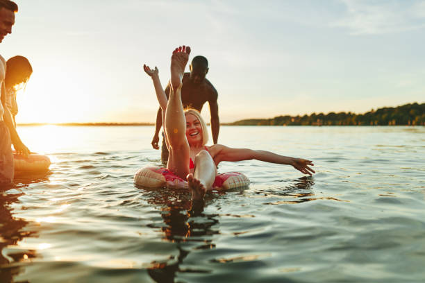 улыбающиеся друзья веселились вместе в озере на закате - inner tube swimming lake water стоковые фото и изображения