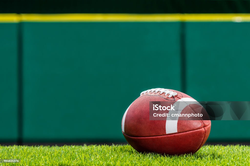 College or high school American Football sitting on the turf of a stadium. A low angle view, of a textured College or High School American Football made of leather with white stripe sitting on artificial turf of a stadium with green padded wall in the background. This type of football with the white stripes is use by colleges and high schools in the US. American Football - Sport Stock Photo