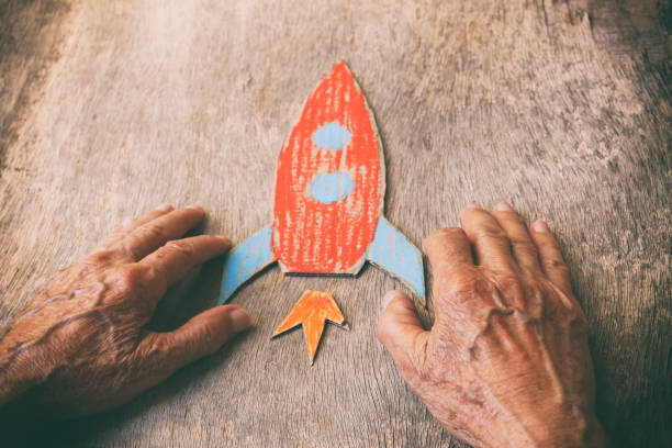 a close up of an elderly man holding a paper rocket on a wooden table. concept of thinking about childhood dreams, sadness and loneliness - distress rocket imagens e fotografias de stock