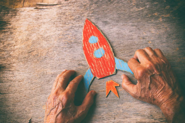 a close up of an elderly man holding a paper rocket on a wooden table. concept of thinking about childhood dreams, sadness and loneliness - distress rocket imagens e fotografias de stock