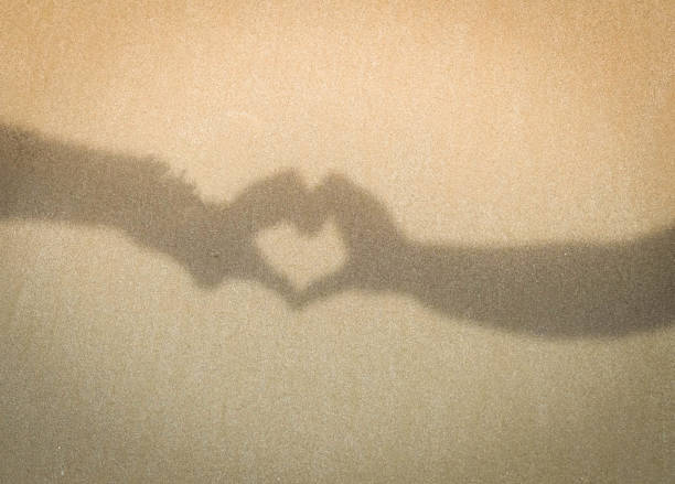 istock Two hands making love heart shadow symbol on the sand at the beach 985884608