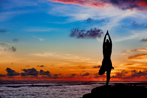 Sunset meditation silhouette. Active woman stand in yoga pose on beach rock to keep fit and health. Healthy lifestyle, fitness training, sport activity on summer family holiday.