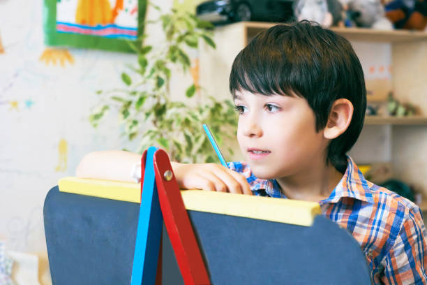 istock Child standingnext to the easel. Kid boy learn paint by brush in class school. Kindergarten interior on background. Boy is getting ready to become an artist 985910488