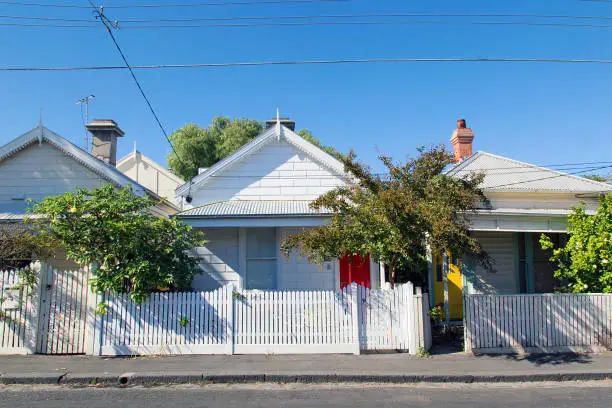 Row of detached bungalow homes in the residential suburb of St Kilda in Melbourne with white picket fence and a blue sky background. Power cables overhead.
