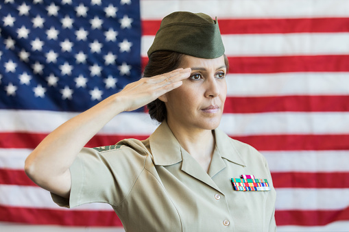 Serious mature Hispanic female military officer in uniform salutes the American flag. She is standing in front of the flag.