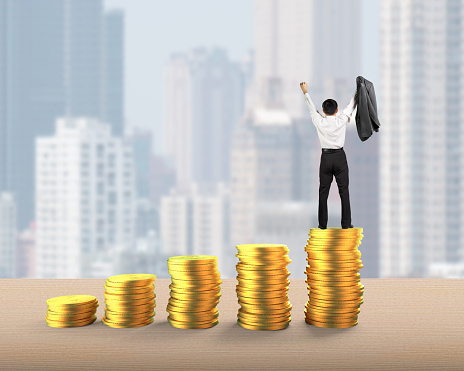 Rear view of businessman standing and cheering on top of golden coins stacks, with city buildings skyscrapers background, concept of business financial growth success.