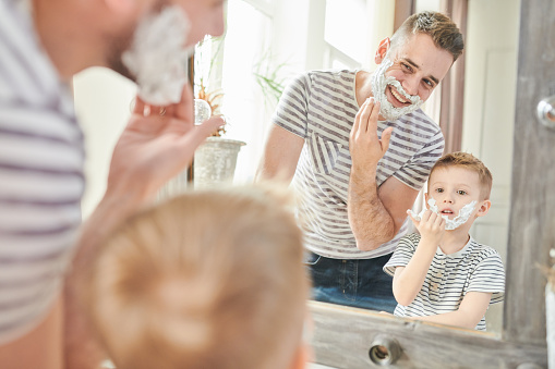 Warm toned portrait of handsome dad teaching cute little son how to shave, standing against mirror with faces covered in foam in modern bathroom