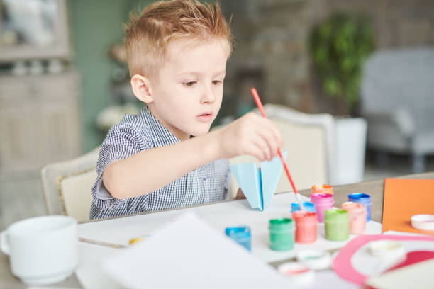 Portrait of cute little boy painting with pastel colors while sitting at kiddies desk in elementary school