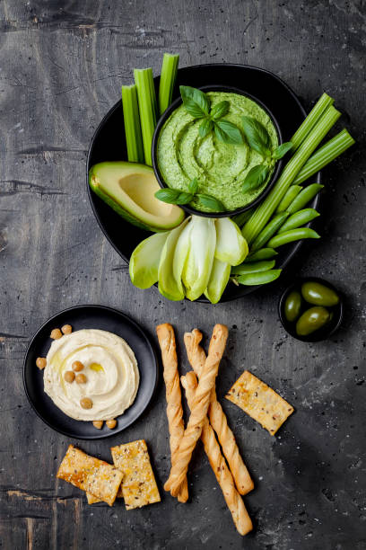 istock Green vegetables snack board with various dips. Hummus, herb hummus or pesto with crackers, grissini bread, fresh vegetables. Healthy raw summer platter. 986091532