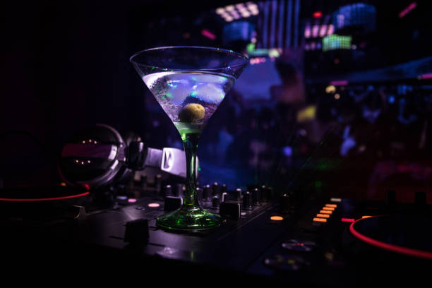 istock Glass with martini with olive inside on dj controller in night club. Dj Console with club drink at music party in nightclub with disco lights. Close up view 986107274