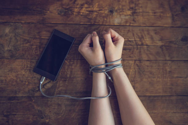 Woman hands trapped and wrapped on wrists with mobile phone cable as handcuffs in smart phone networking and communication technology addiction concept Cropped shot of woman with hands tied with smartphone wire, phone addiction concept. woman hands with phone on wooden background smart phone technology lifestyles chain stock pictures, royalty-free photos & images