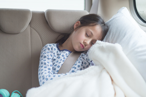 An elementary-age ethnic girl sleeps in the back seat on a family road trip. She is resting her head on a pillow.