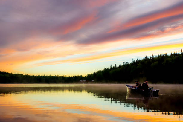 Two fishermen on a peaceful lake with a multicoloured sky sunset and a chalet in background. Quebec, Canada