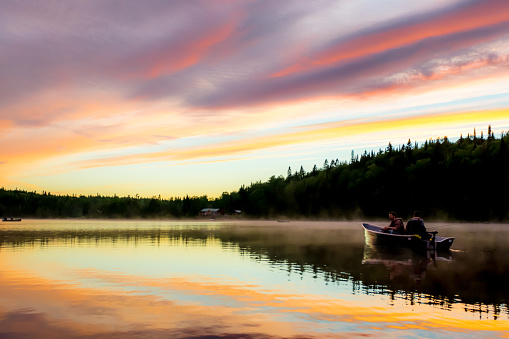 Two fishermen on a peaceful lake with a multicoloured sky sunset and a chalet in background. Quebec, Canada