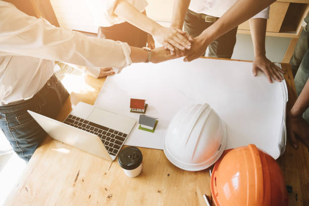 istock Young colleagues joined together their hands, Support together with blueprint of construction project ,small house model and safety helmet in office, Construction Concept. 986342340