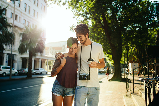 Happy explorer couple walking in the street holding a coffee cup with sun in the background. Man carrying a camera and holding a coffee cup walking with a woman with his arm around her shoulders.
