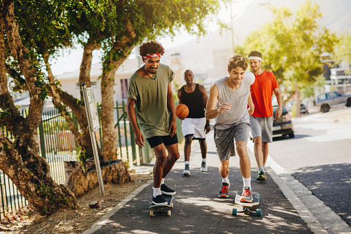 Two men skating on skateboard on a pavement while their mates cheer them. Basketball guys walking on pavement using skateboards.