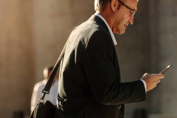istock Senior man using mobile phone while walking on street to office 986481452
