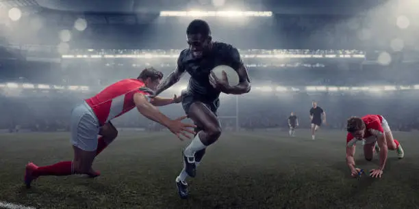 A mid action image of a professional black male rugby player running whilst holding a rugby ball and running pushing away a rival player who is about to tackle. The athlete has mouth open and teeth bared in determination. The action occurs in a generic misty floodlit rugby stadium.