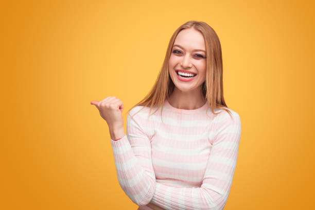 Laughing young woman with makeup wearing casual striped shirt and pointing away with thumb looking at camera on orange background