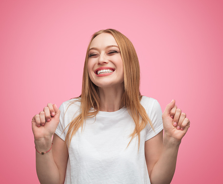Smiling casual blond woman in white t-shirt holding hands up in excitement and anticipation while keeping eyes closed on pink background