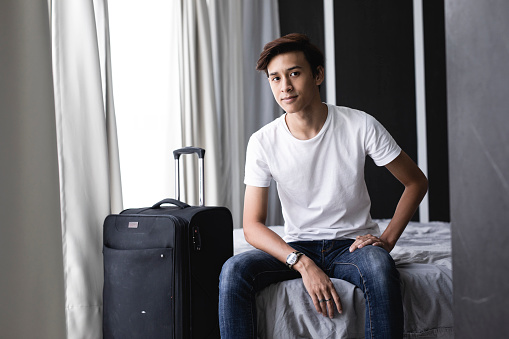 Young asian boy sitting on a bed in a hotel room with a suitcase.