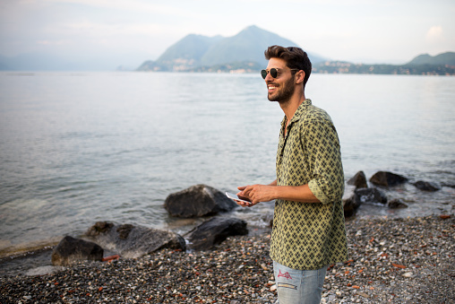Handsome smiling man wearing sunglasses and looking away. He is walking by the sea and using mobile phone