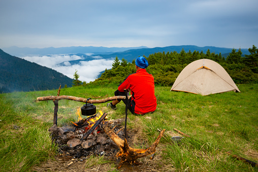 Adventurer sits in the camp next to the bonfire on a green meadow and admires the mountain ranges in the fog after the storm. Epic travel in the mountains. Back view.