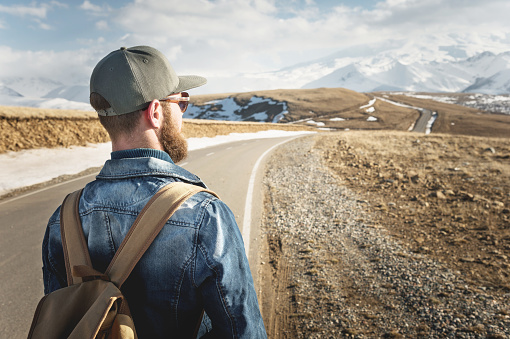 A bearded man in a cap with a backpack ready to go a long way. A man on a country road against the backdrop of mountains and clouds.