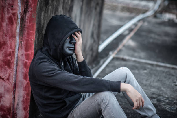 istock Mystery man with black mask in headache gesture feeling stressed and depressed while sitting near grunge concrete wall in abandoned building 986650318