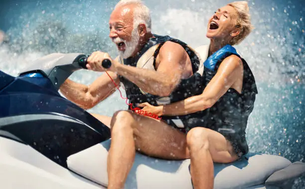 Closeup side view of a senior couple riding a jet ski on a sunny summer day at open sea. The man is driving quickly through the waves, and the lady is hardly holding on. Caught in the moment of max speed.