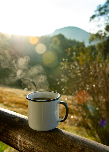 White enameled cup with hot coffee, steaming out on the wooden fence in the farm house