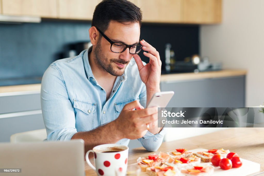 Breakfast Attractive young man in denim shirt typing message on his phone while having breakfast at home Healthy Lifestyle Stock Photo