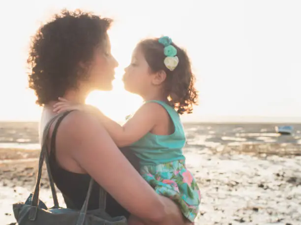 Little girl and her mom kissing on the beach during sunset. In the background you can see the wet sand due to the low tide. Vintage filter with soft style