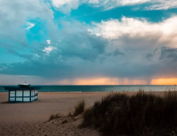 beach hut at sunset on the sand of the beach, storm clouds are seen in the horizon unloading on the sea water, between which sunlight filters. In the foreground vegetation are seen