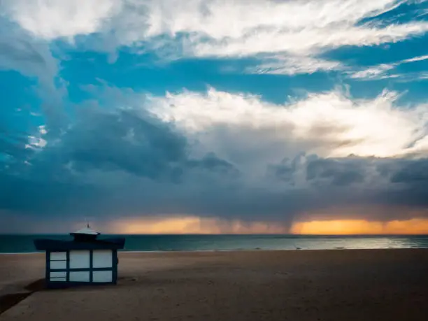 beach hut at sunset on the sand of the beach, storm clouds are seen in the horizon unloading on the sea water, between which sunlight filters.