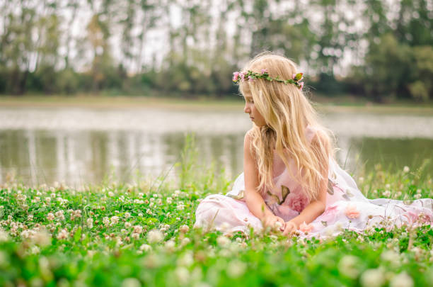 sad lonely girl with long blond hair in pink dress sitting alone as the fairy in the green grass by the river