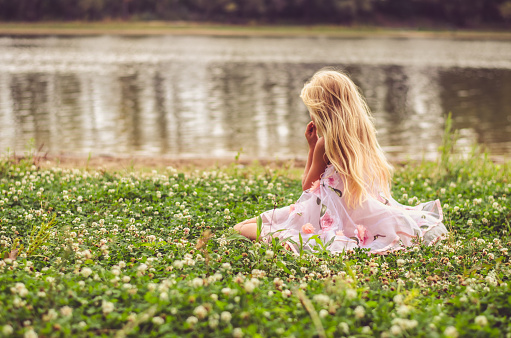 sad lonely girl with long blond hair in pink dress sitting alone as the fairy in the green grass by the river