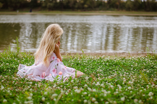 sad lonely girl with long blond hair in pink dress sitting back view alone as the fairy in the green grass by the river